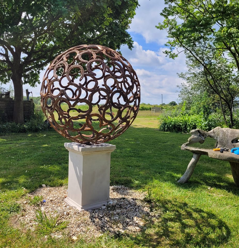 Metal sphere sculpture on plinth within large garden
